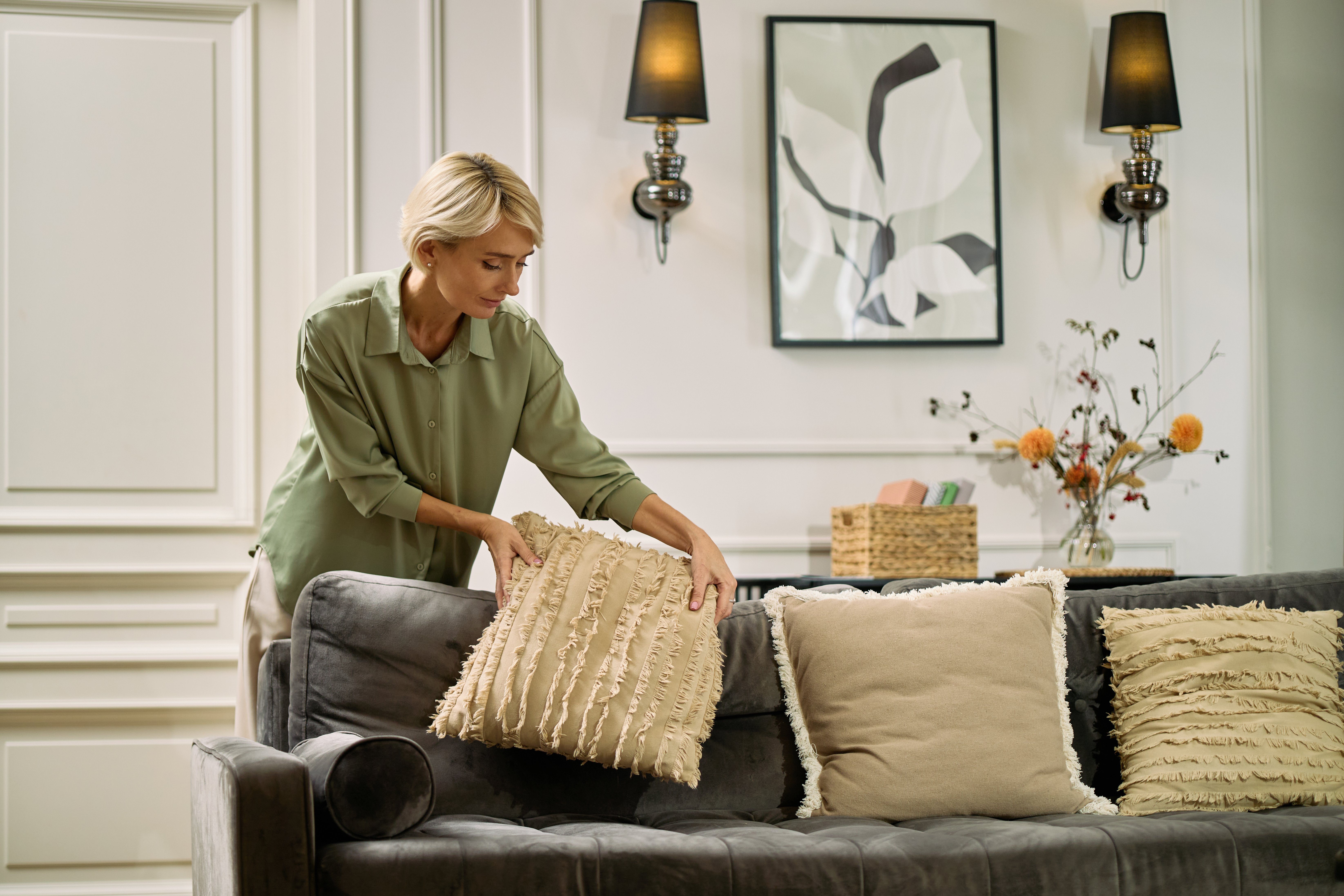 Caucasian middle aged woman arranging decorative pillows on sofa in modern living room, standing and leaning forward while focusing on organizing cushions in comfortable home setting