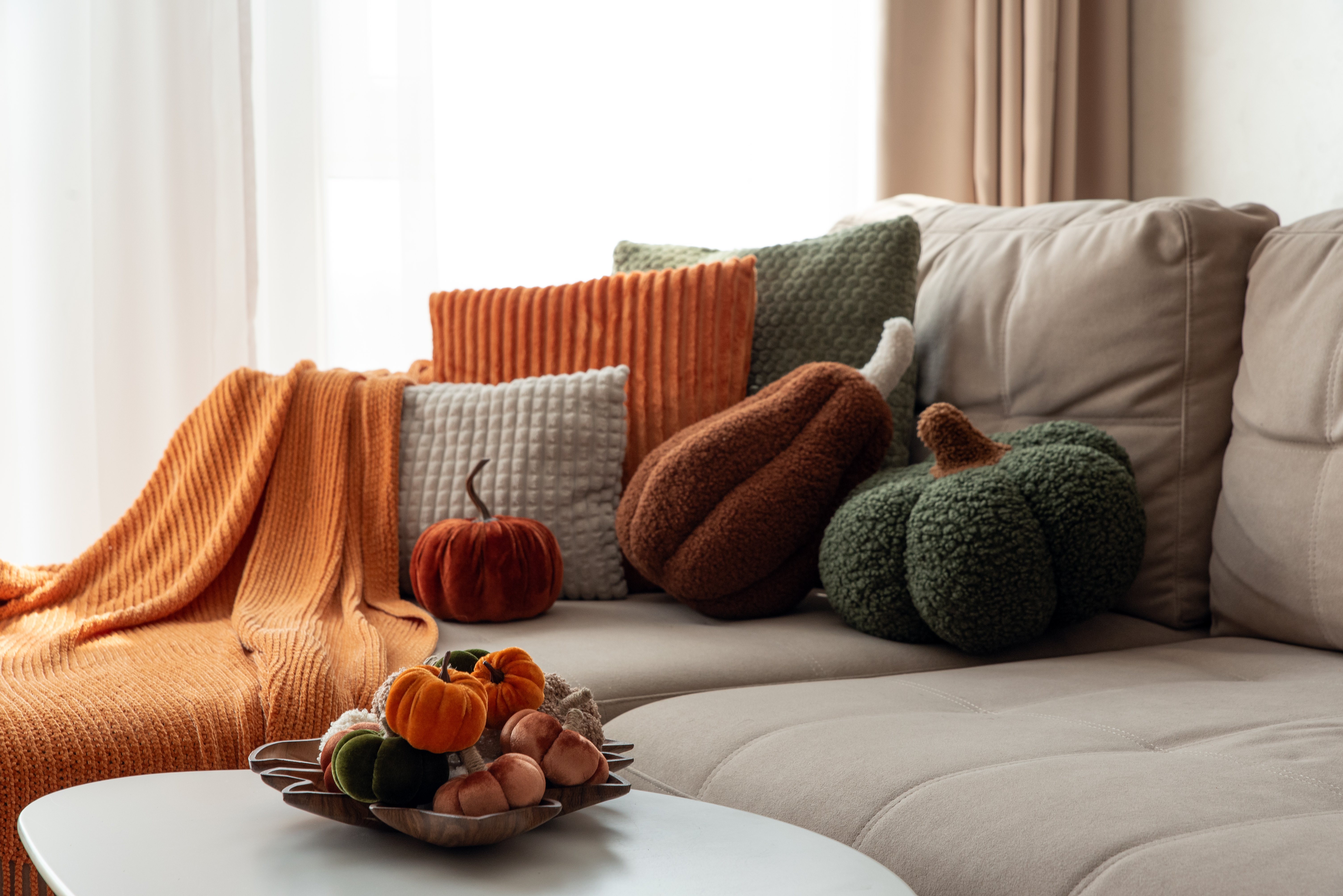 Soft lighting illuminates plush textured pillows and a chunky knit throw on a neutral sofa, accented by an assortment of decorative velvet pumpkins. Cozy home decor suggesting harvest season and hygge